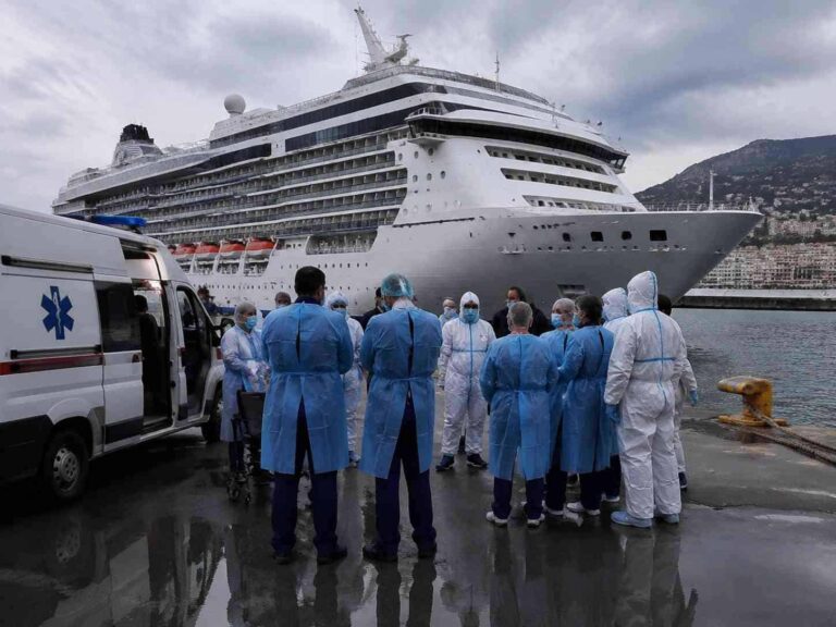 Medical team in blue gowns and white protective suits stand in a circle near a large cruise ship at a harbor, with an ambulance visible nearby.