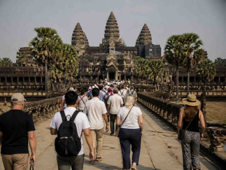 Tourists walk along a stone causeway toward a Khmer temple with three central towers, flanked by palm trees on both sides.