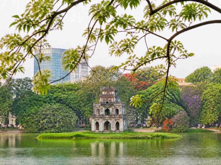 Three-story stone pagoda on a small island in a calm lake, framed by surrounding trees with a cityscape in the background.