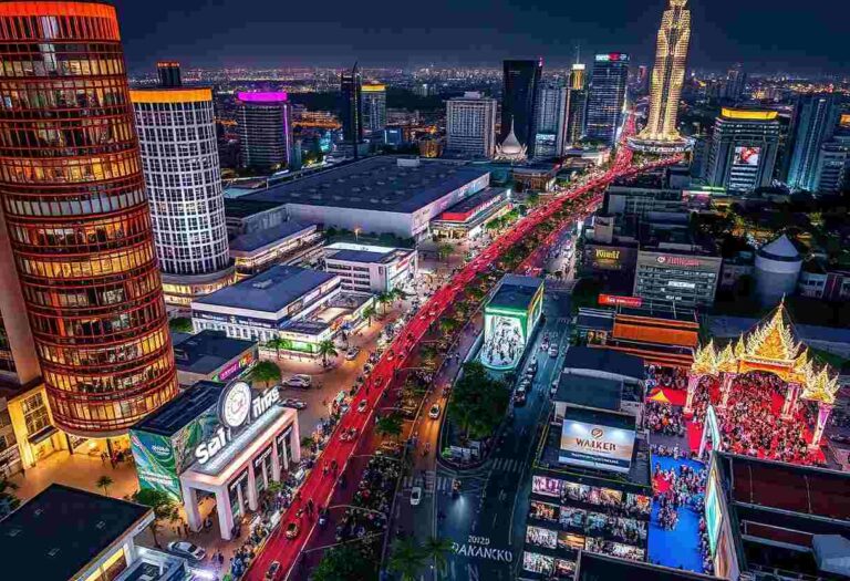 Aerial night view of a busy city street lined with neon-lit buildings and a crowded festival area on the right side.