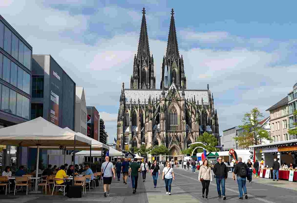 Streetside festival with outdoor tents and cafe seating leading to the Cologne Cathedral's twin spires in the background,