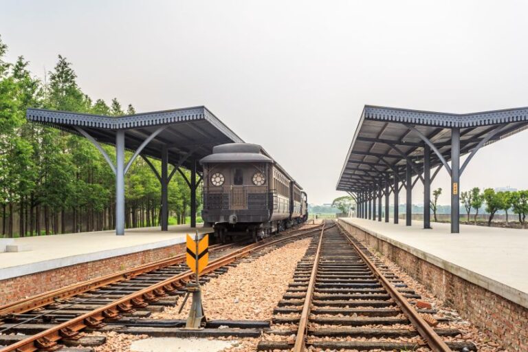 Vintage steam locomotive on tracks between two covered platforms at a quiet railway station, with green trees in the background.