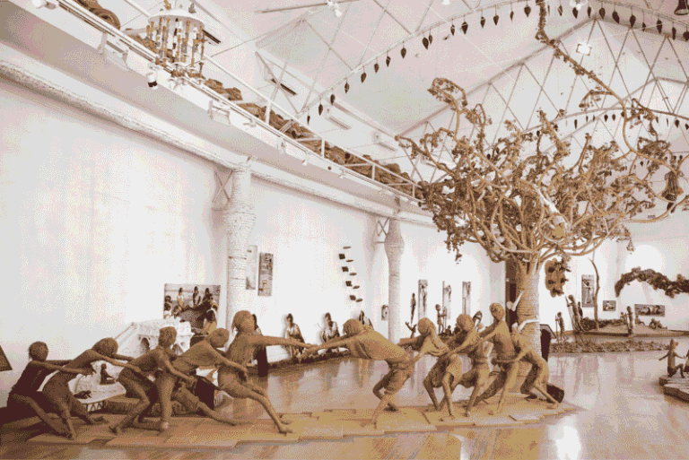 People in a bright, spacious studio engage in a tug-of-war-style team exercise as a large sculpted tree stands in the background on the right.