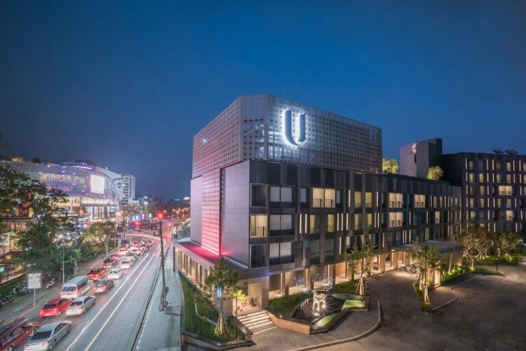 Nighttime view of a modern mixed-use building with a perforated metal facade and a large illuminated U logo atop the structure near a busy street.