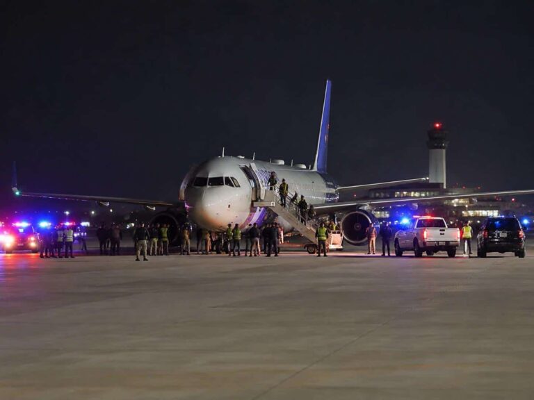 Night-time scene of a large passenger jet on the tarmac with passengers boarding via mobile stairs as emergency/ground vehicles with flashing lights surround the plane.
