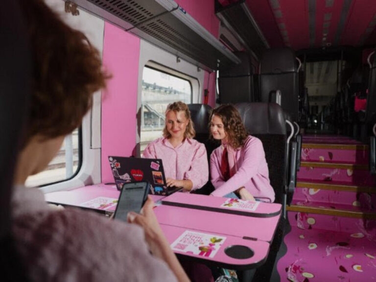 Two women in pink sit at a pink-trimmed train table, looking at a laptop together.