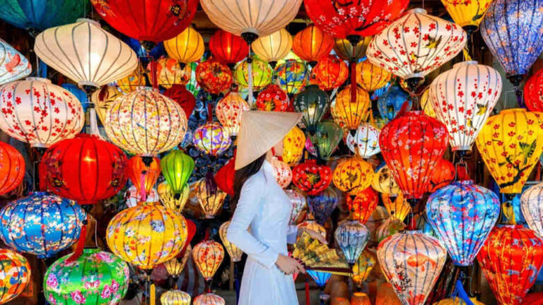 Woman in a light blue ao dai and conical hat among a rainbow of colorful lanterns at a festival market�ting scene.
