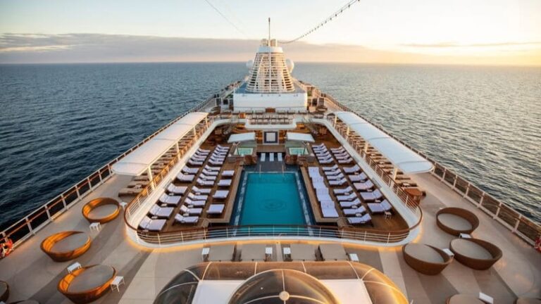 Aerial view of a cruise ship's pool deck with a central rectangular pool, rows of white lounge chairs, and circular seating areas along the sides at sunset.