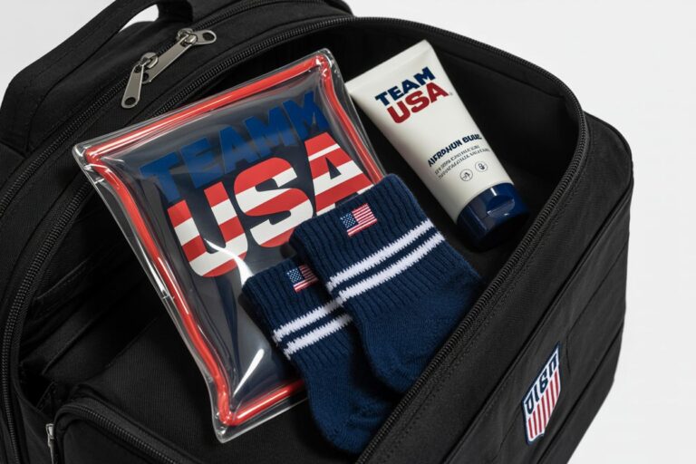 Open black backpack with Team USA gear: a red‑blue zipper pouch, navy striped socks, and a white Team USA tube inside.