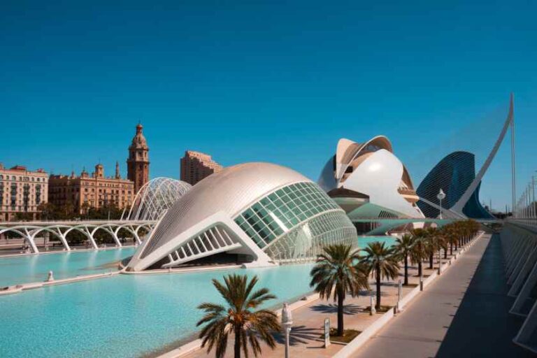 Futuristic white buildings beside a reflective water pool, palm trees lining a promenade under a clear blue sky (Valencia’s City of Arts and Sciences)