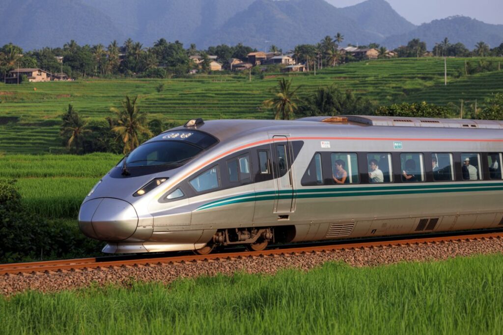 Silver high-speed train gliding along a single track through green fields and distant mountains in the background.