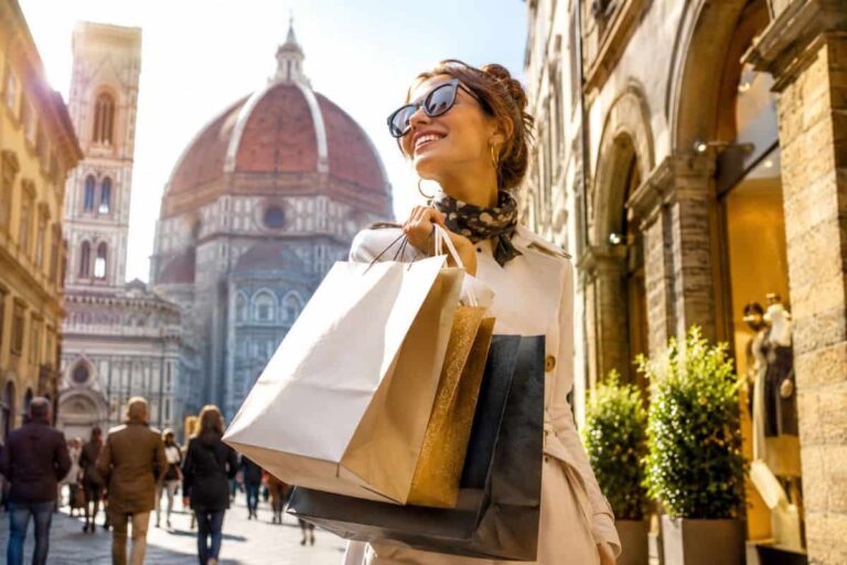Woman smiling with several shopping bags in a sunny Italian street near a historic cathedral dome.