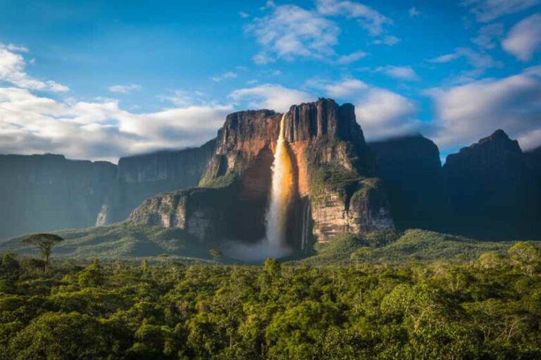 Waterfall cascading down a towering cliff into a dense tropical jungle under a blue sky.