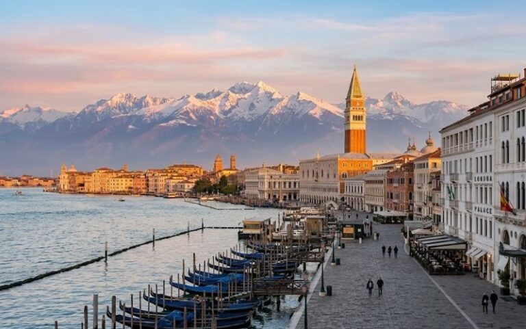 Venice waterfront with rows of gondolas along a stone quay and a bell tower, with snow‑capped mountains rising in the distance at sunset.