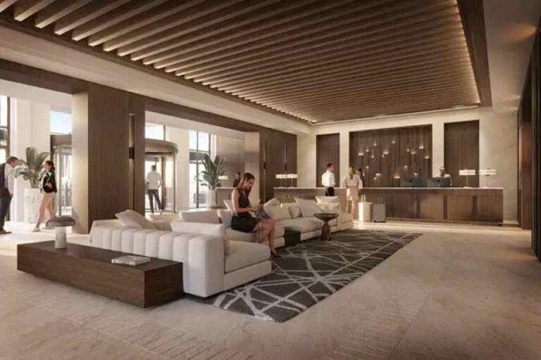 Modern hotel lobby with white sectional sofas, a patterned rug, and a front desk in the background where staff assist guests.
