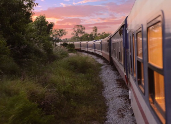 Passenger train curving along a gravel track beside green bushes at sunset, windows reflecting warm light.
