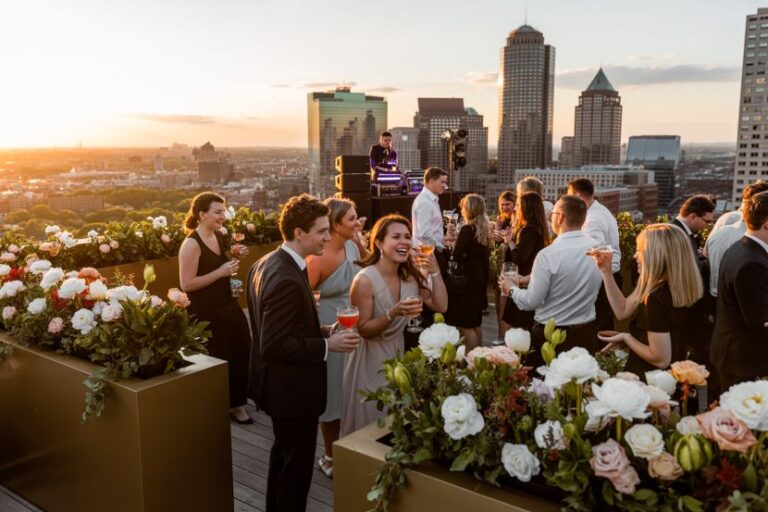 Group of guests in formal attire mingling on a rooftop at sunset, holding drinks, with large flower planters and a city skyline in the background.