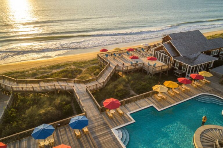Aerial view of a beachfront resort with a wooden deck, pool, and colorful umbrellas along the shore.