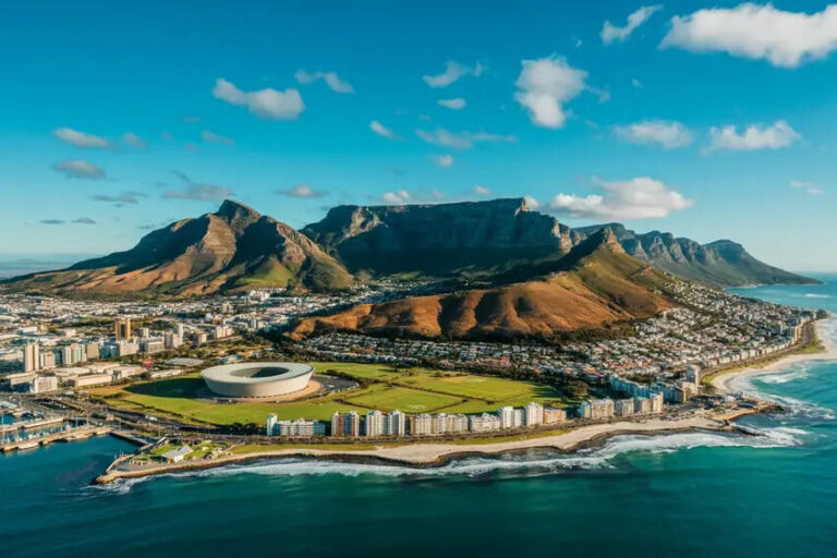 Aerial view of Cape Town area showing Table Mountain, a circular stadium, and a green waterfront district by the sea.