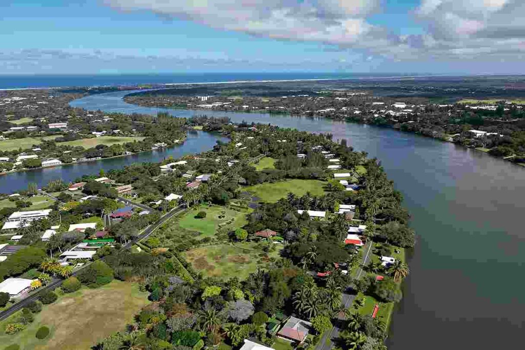 Aerial view of a coastal town along a winding river with green yards and palm trees.