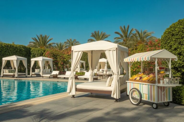 Poolside resort scene with white cabanas, loungers and a striped fruit cart by the blue pool under palm trees on a sunny day.