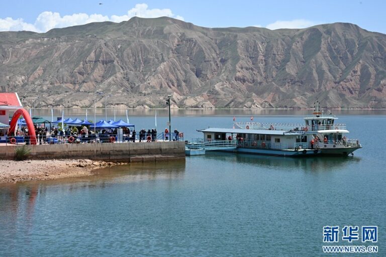 People stand along a lakefront promenade under blue canopies while a white tour boat is docked at the pier, with rugged mountains in the background on a sunny day.