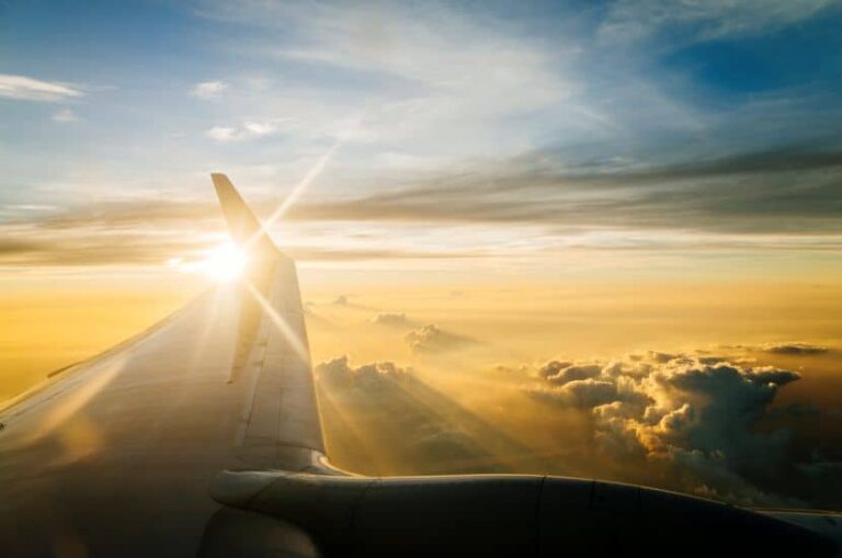 Airplane wing with sun bursting through clouds during sunrise above golden skies.