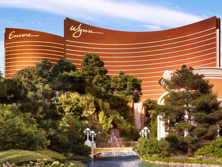 Facade of the Encore at Wynn Las Vegas with curved orange walls and white lines, surrounded by lush trees and a water feature in the foreground.