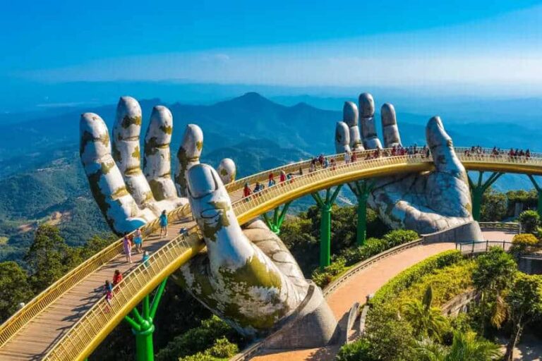 Giant stone hands cradle a curved pedestrian bridge with tourists walking along, set in a mountainous landscape.