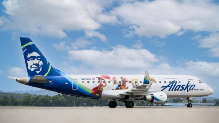 Alaska Airlines jet parked on the tarmac with a colorful mural of cartoon characters along the fuselage and a large portrait on the tail fin