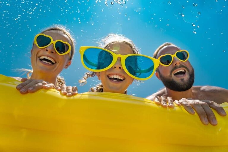 Three friends with bright yellow sunglasses grin over a yellow inflatable raft in a sunny blue pool/sea.