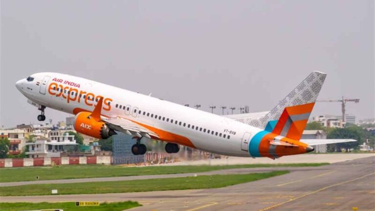 Air India Express passenger jet lifting off from a runway with orange and blue livery, city buildings in the background.