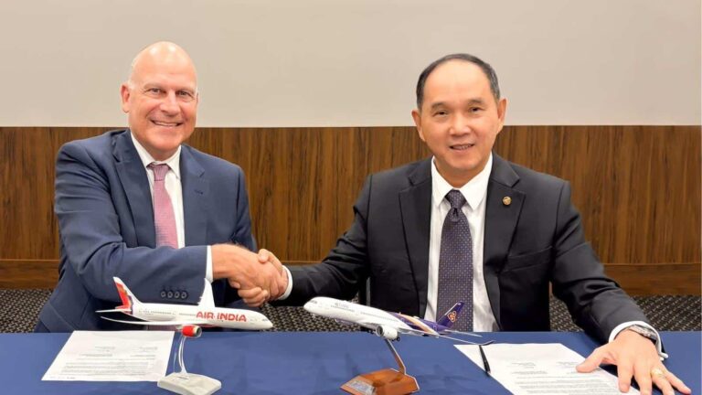 Two businessmen in suits shake hands across a signing table, with two miniature airplanes (including an Air India model) and documents in front of them, signaling a partnership signing.