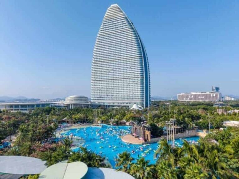 Tall sail-shaped hotel (Burj Al Arab) behind a large blue lagoon-style pool and palm trees on a sunny coast.