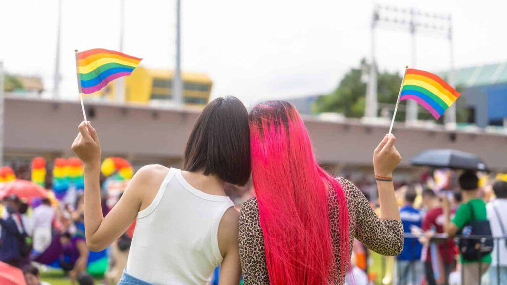 Two people stand with their backs to the camera, waving rainbow Pride flags at a lively outdoor event. Two friends hug or lean together, each holding a rainbow flag, with a blurred crowd in the background.