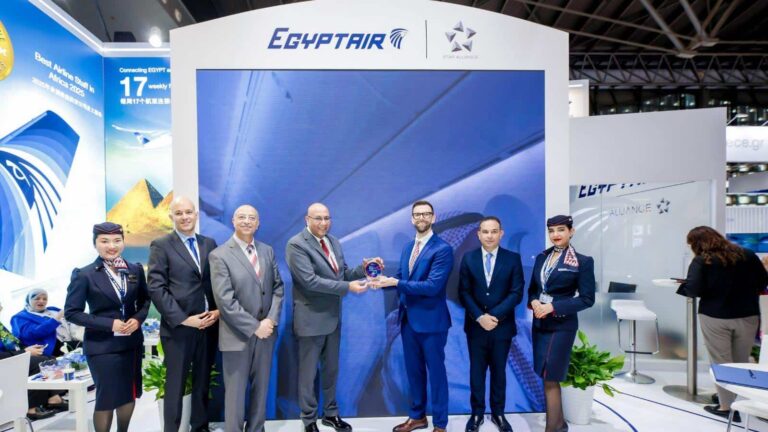 Eight professionals in suits (two women in uniform) stand at an EgyptAir booth as one man hands a glass award to another in front of a blue backdrop with logos.