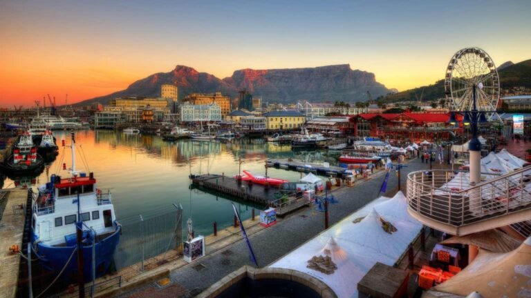 Harbor at sunset with boats, ferris wheel, and colorful buildings against distant mountains (Table Mountain). **Cape Town waterfront.**