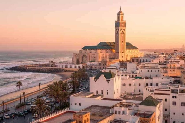 Coastal city at sunset with a large mosque and tall minaret overlooking the sea and white buildings along the shore.