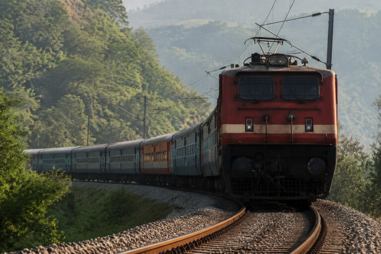 Electric red locomotive leading a long blue passenger train along a curved track through green countryside and hills behind it.