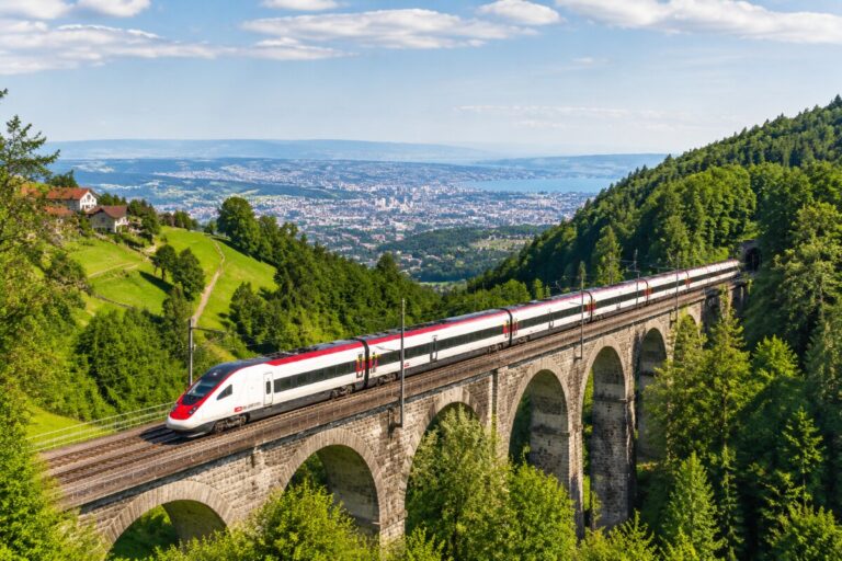 Modern white and red high-speed train crossing a stone-arched viaduct over a green valley with a distant cityscape.