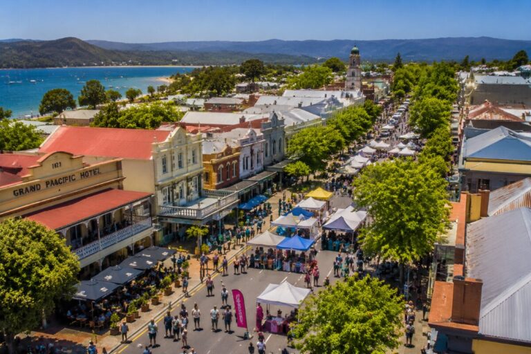 A sunny seaside street market with rows of tents and crowds, along a waterfront town with historic buildings and green trees.