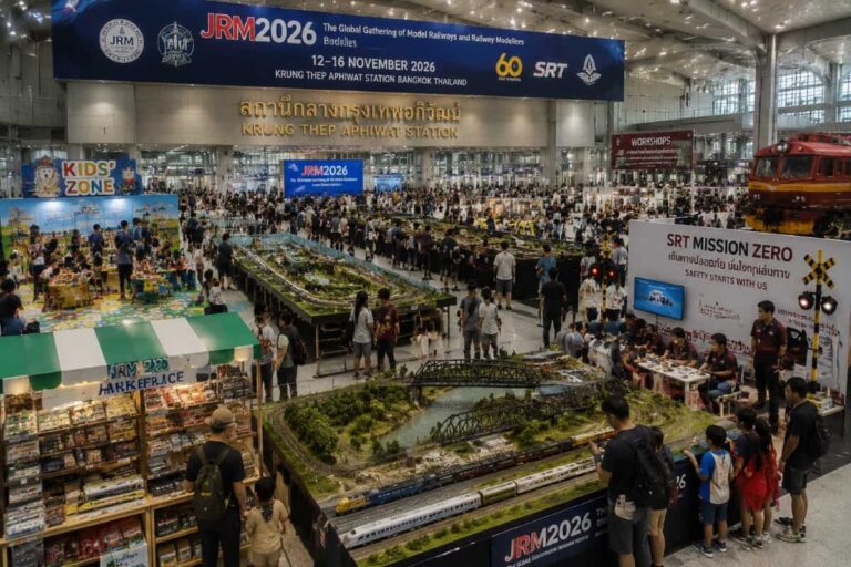 Busy model railway exhibition hall with detailed miniature train layouts, stalls, and visitors entering the showfloor in Bangkok