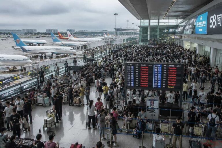 Crowded airport terminal concourse with many passengers waiting with luggage; an electronic board shows flights marked as Cancelled or delayed and planes are visible on the tarmac outside.