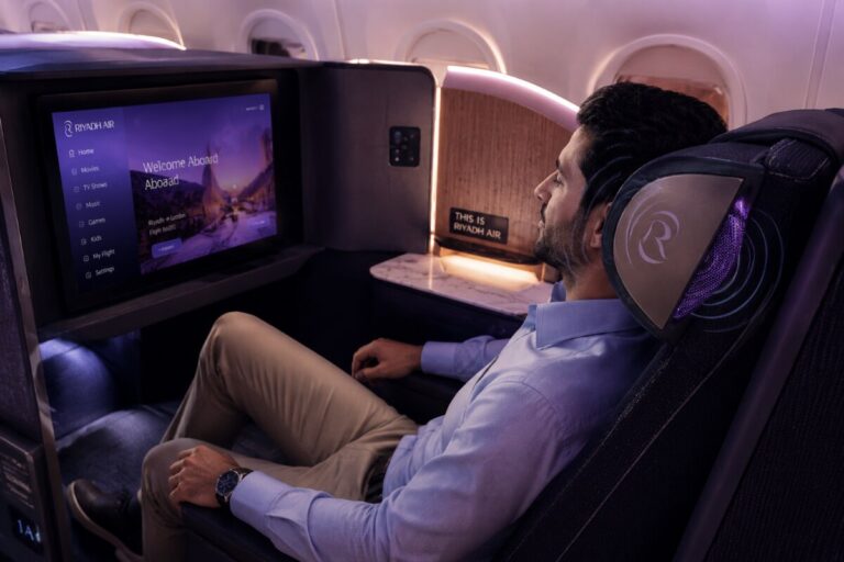 Passenger in a premium cabin seat with headphones, watching the in-flight entertainment screen showing ‘Welcome Aboard’ and Riyadh Air branding in a dimly lit cabin.