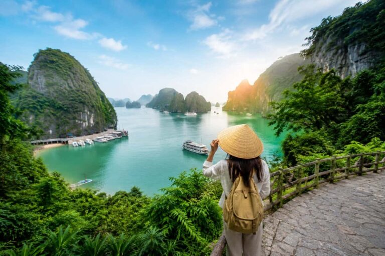 Traveler with a straw hat and tan backpack stands on a stone path overlooking emerald bay with limestone karsts and boats in Ha Long-like scenery at sunset