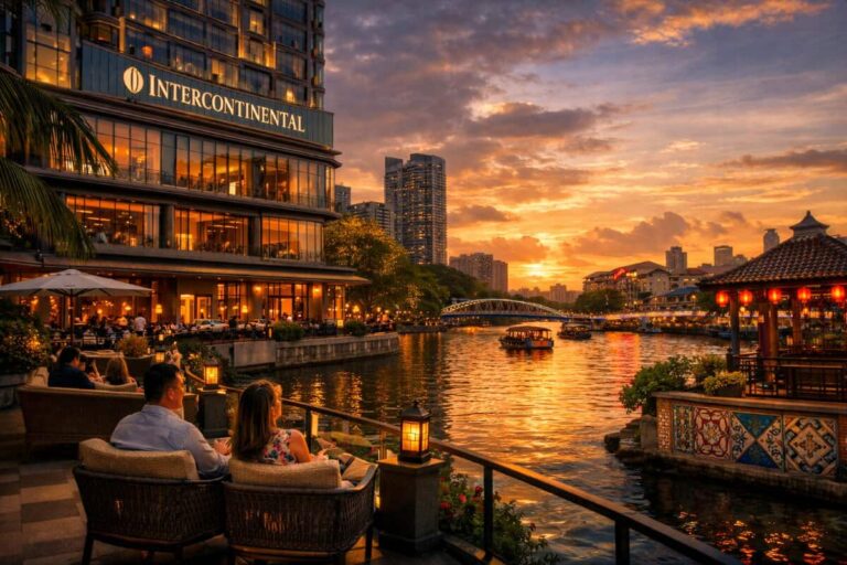 People dining on a riverside terrace at sunset, with an illuminated Intercontinental hotel and a waterway park scene in the background