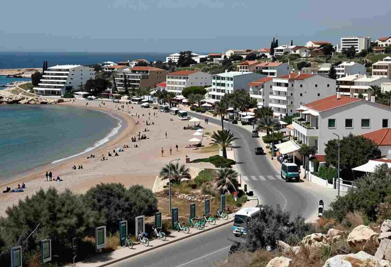 Coastal town with a sandy beach, turquoise water, and white multi-story buildings along a curved road.