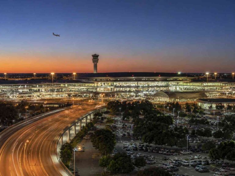 Sunset over a busy airport with a control tower, glass terminal, and a curving highway in the foreground; plane in the sky.