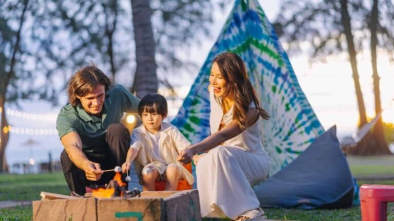 Family of three roasting marshmallows over a campfire beside a colorful tie-dye tent in a park at sunset. Their smiles show a cozy outdoor camping moment.