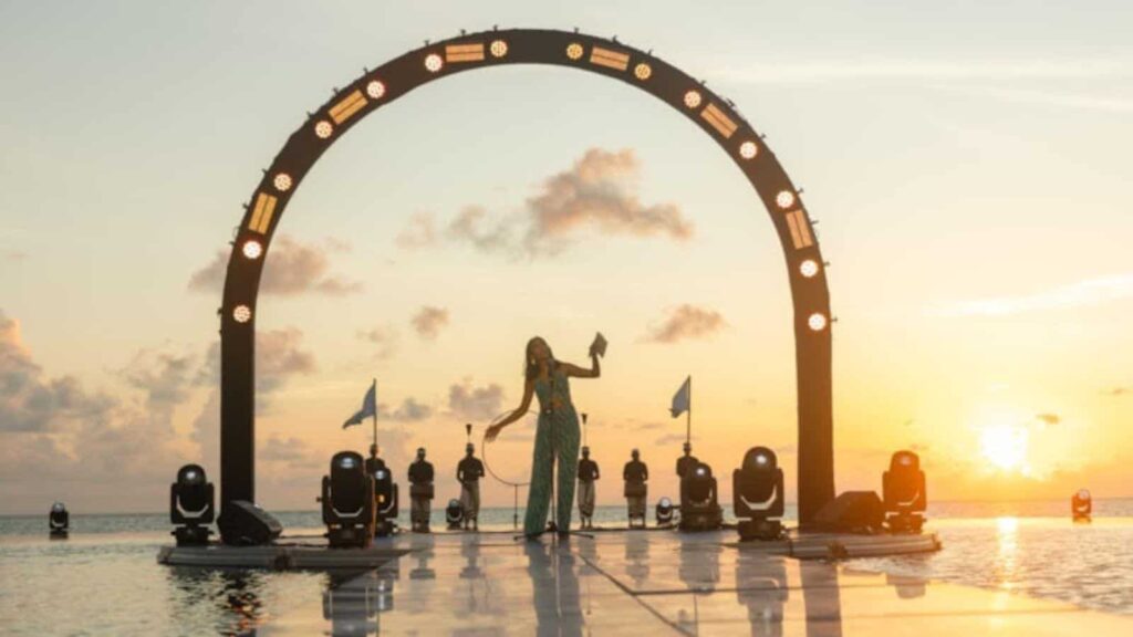 Female dancer in a teal gown performs on a reflective water stage beneath a large illuminated semicircular arch at sunset.